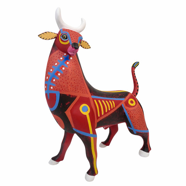 A hand-carved , hand-painted Mexican copal wood sculpture of a bull, featuring a vibrant red and black body with geometric patterns in yellow, blue, and white, set against a plain white background.
