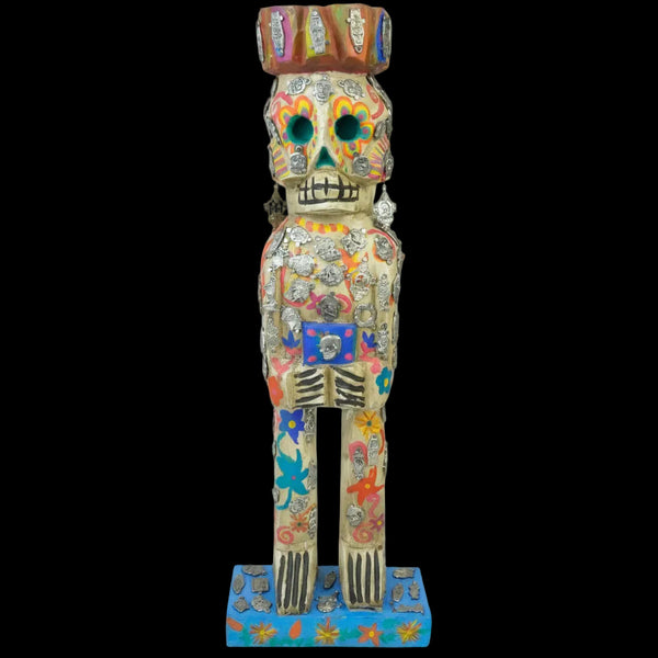 A colorful Day of the Dead (Dia de los Muertos) sugar skull wooden figurine decorated with silver skulls and milagros, standing on a blue patterned base against a white background.