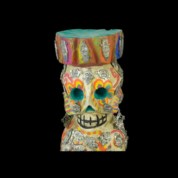 Colorful skull-shaped wood carving with decorative milagros on a white background