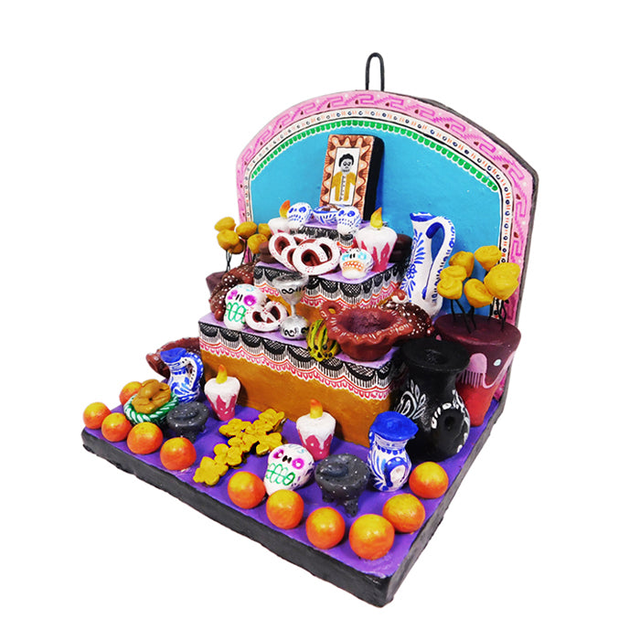 A colorful Day of the Dead ceramic ofrenda featuring sugar skulls, day of the dead bread, bananas, liquor, a molcajete, and flowers on a purple base.